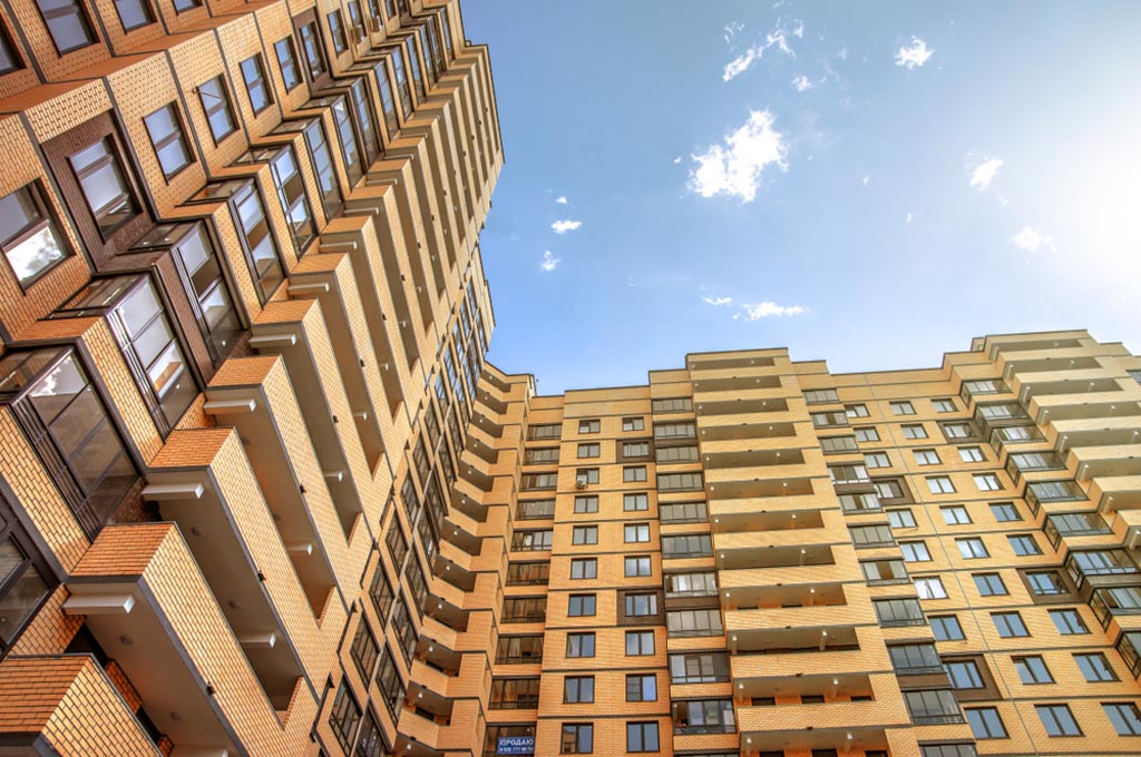 alt="A multi-story modern apartment building with numerous balconies and windows set against a blue sky, requiring intumescent fire protection to increase the fire resistance of structural steel beams."